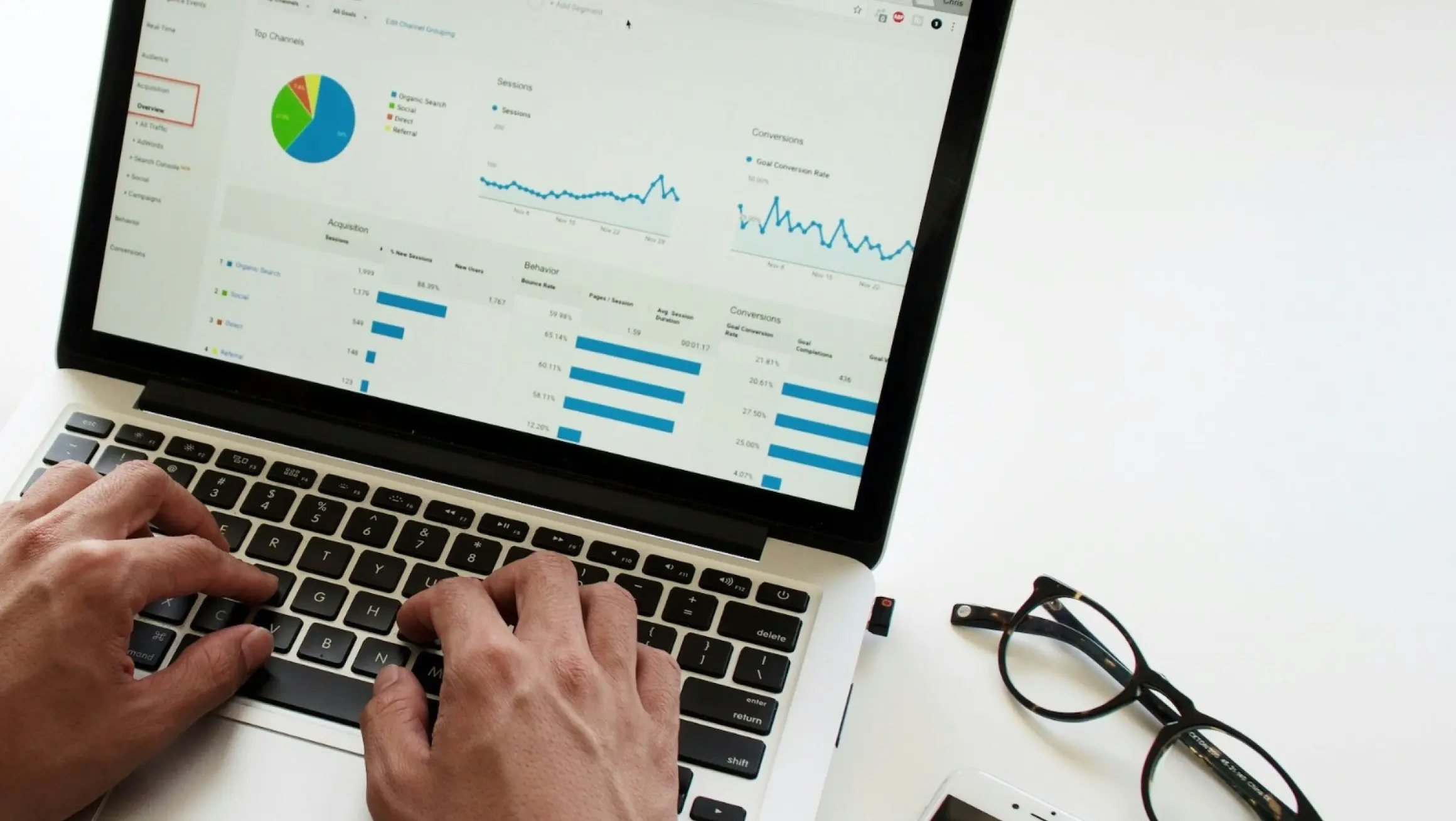 Hands typing on a laptop keyboard with a screen displaying various graphs and charts related to data analytics. Nearby on the white surface are a pair of black-rimmed glasses and a smartphone.